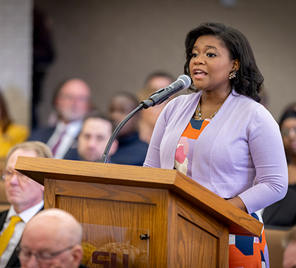 Alcina standing at a lectern speaking in a crowd of people.