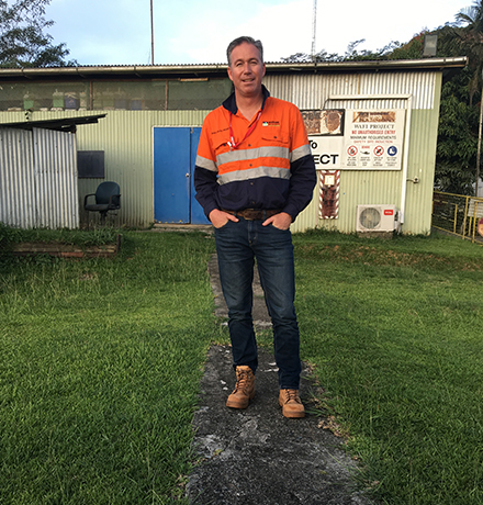 David wearing an orange high-visibility work shirt, jeans, and brown work boots standing on a narrow concrete path outside an industrial building with safety signage