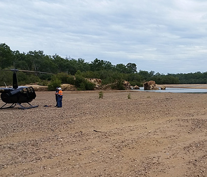Iain standing next to Helicopter on dry riverbed near water