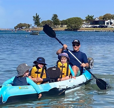 Stuart in a boat with 3 children paddling with oars.