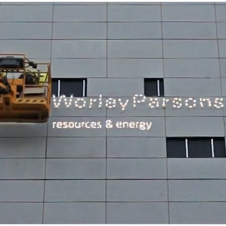 Animated image of a person in a crane installing WorleyParsons logo on the side of a building.