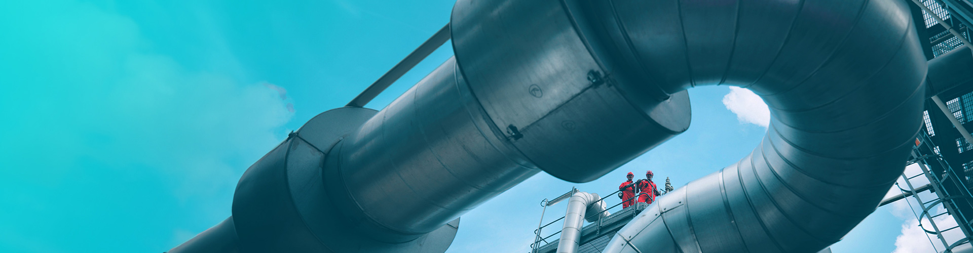Looking upward at two people standing on a platform, past large steel pipes at a plant,