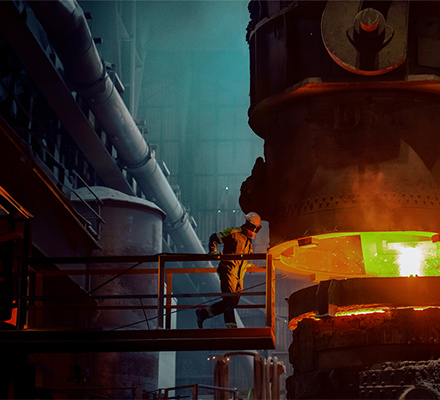 Worker wearing protective gear pours molten metal from a ladle into a mould in an industrial facility.