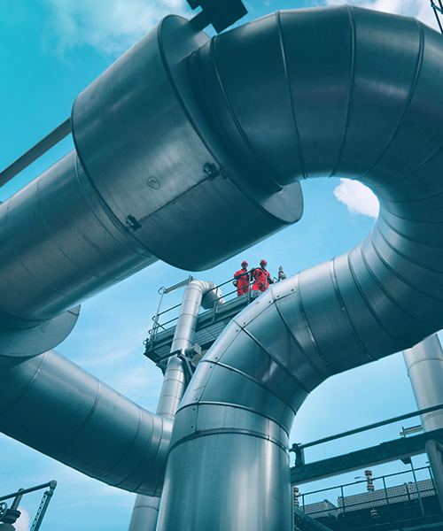 Looking upward at two people standing on a platform, past large steel pipes at a plant,