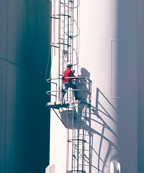 Worker on a ladder attached to the side of a large white tank at a plant.