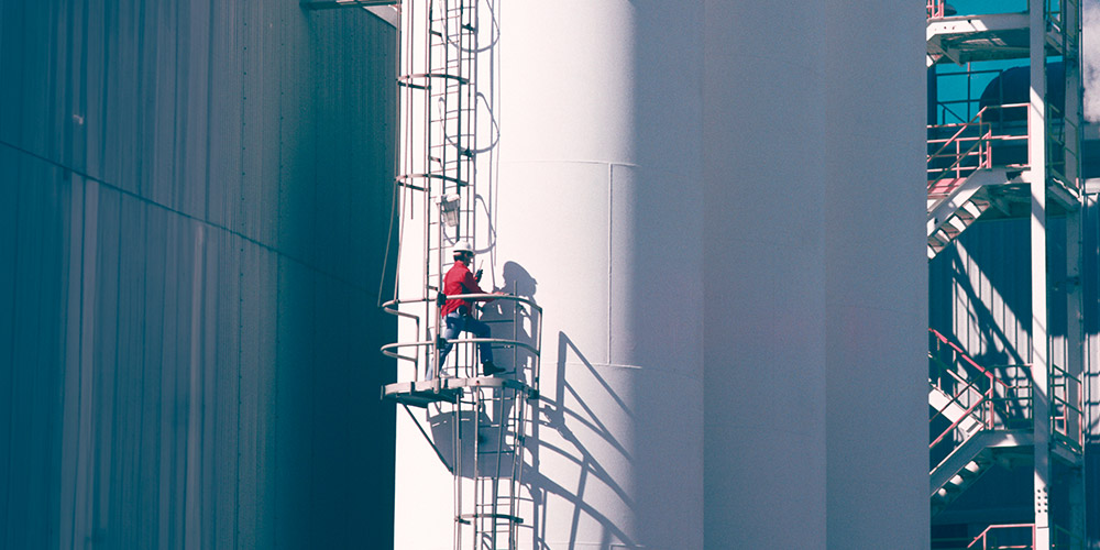 Worker on a ladder attached to the side of a large white tank at a plant.
