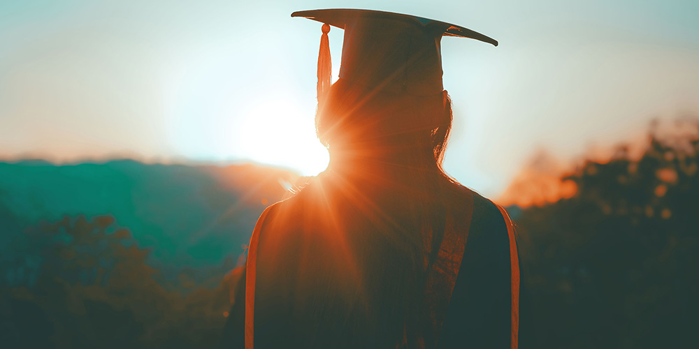 Person wearing graduation gown and cap in front of a bright sun.