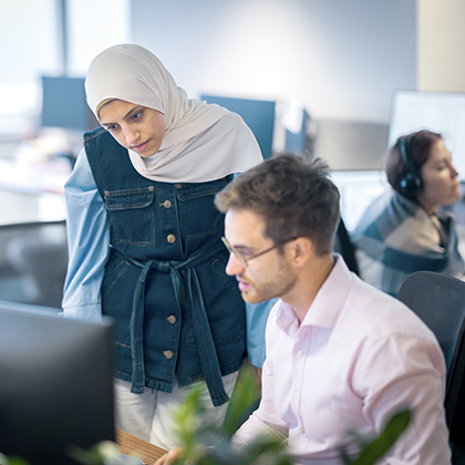 In Worley office, woman standing next to a young man at a desk both looking at the computer.