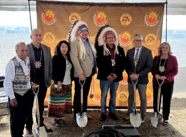 Group of seven people together holding shovels, two men wearing Indigenous headdress.