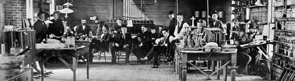 Early 20th-century laboratory with people working at benches filled with electrical equipment.