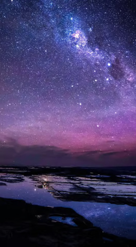 Star-filled night sky with the Milky Way visible above a rocky shoreline and tidal pools.
