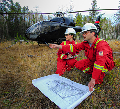 Two people wearing PPE with a geo map crouching in front of a helicopter.